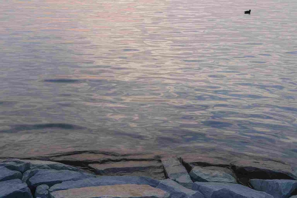 Calm water surface reflecting sunset colors near a rocky shore, creating a peaceful and meditative scene.